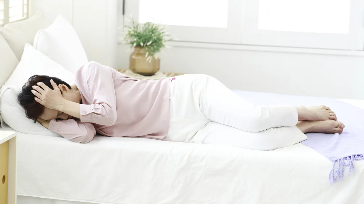 침대에 옆으로 누워 머리를 감싸 쥔 여성, 어지럼과 무기력감을 표현한 장면